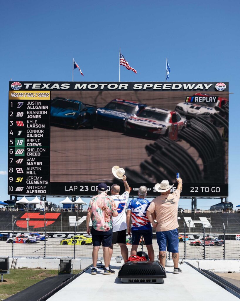 Four fans raise a hat and a drink toward the speedway’s giant screen.