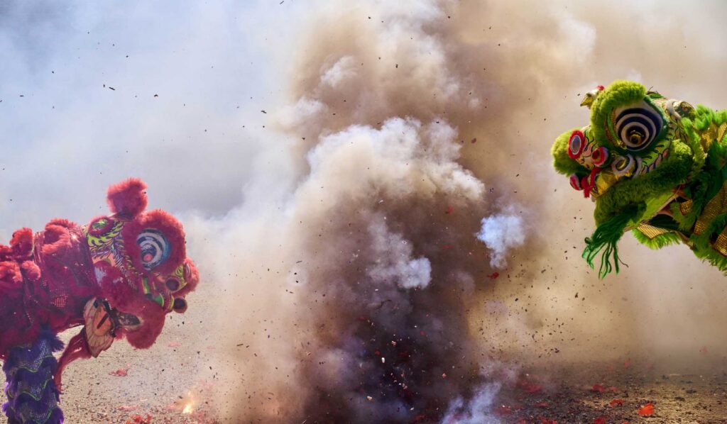 Red and green lions move through smoke and exploding firecrackers.