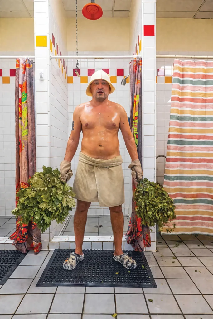 A man in a towel and Crocs at the Russian Banya holds a bushel of wet oak leaves.