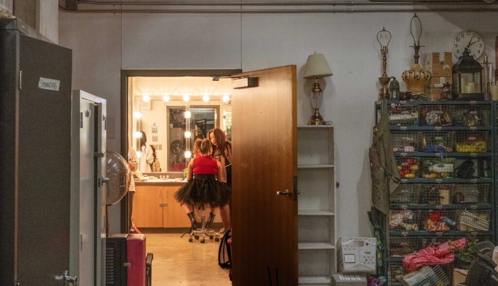 Students prepare in a lit dressing room beyond backstage storage.